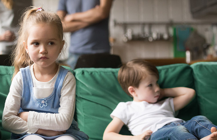 Siblings unhappy and fighting sitting on couch with parents in background - feature