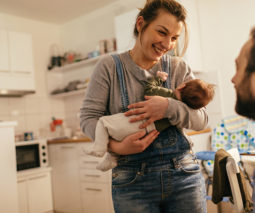 Mother holding newborn talking to partner in kitchen - feature