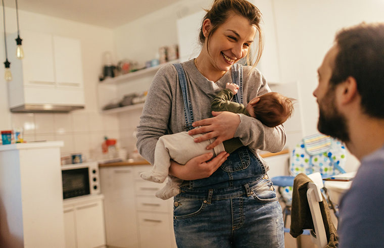 Mother holding newborn talking to partner in kitchen - feature
