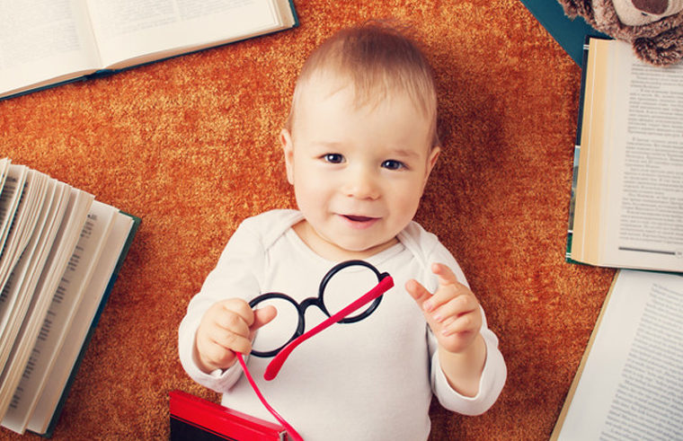 Cute baby boy holding a pair of glasses, surrounded by books