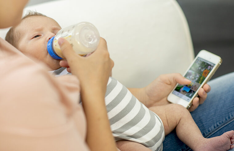 Mother bottle feeding newborn infant while scrolling on phone