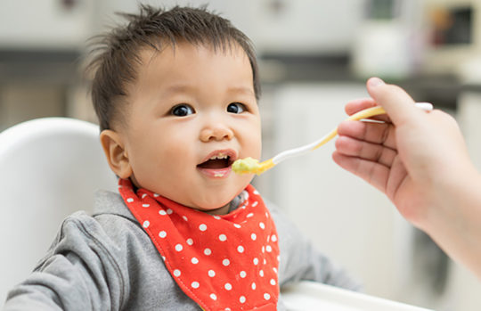 Asian baby sitting in highchair eating from spoon - feature