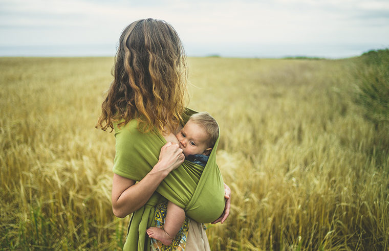 Mother breastfeeding baby in a sling standing in a field - feature