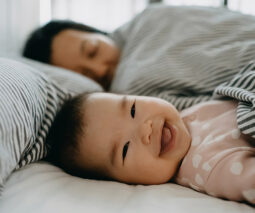 Close up of baby smiling happily on bed while mother sleeping besides her