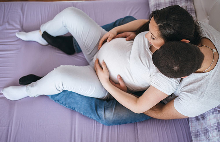 Pregnant couple sitting on bed holding belly -feature