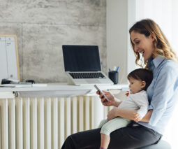 Mother working from home with baby in lap with mobile phone and laptop - feature
