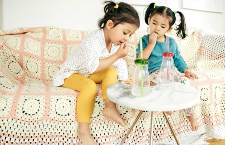 Two young girls sitting on couch eating snacks - feature