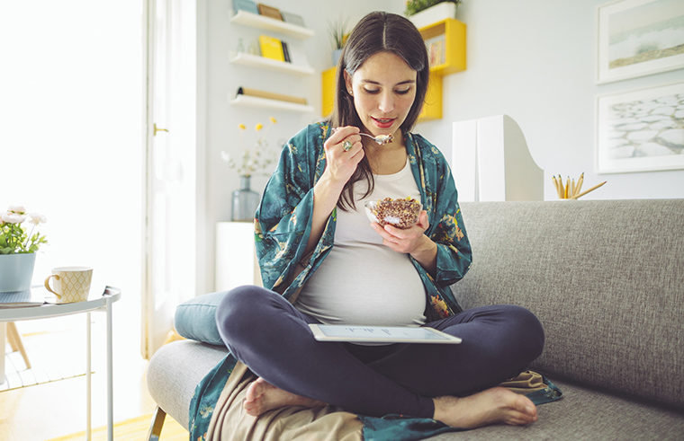 Pregnant woman on couch eating looking at ipad - feature