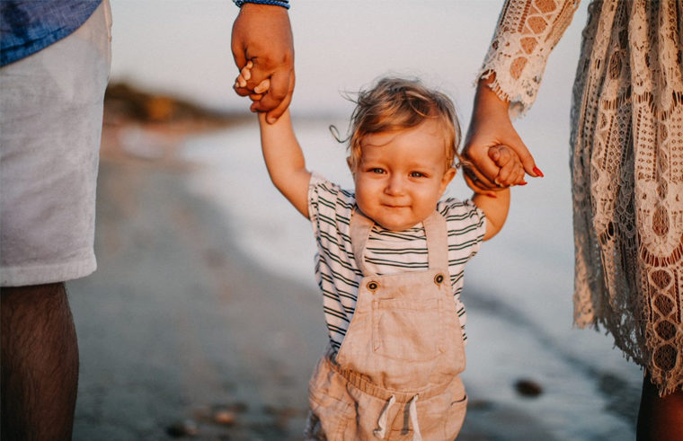 Toddler boy walking on beach holding parents' hands - feature