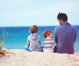 Father sitting on sand at beach with two children - feature