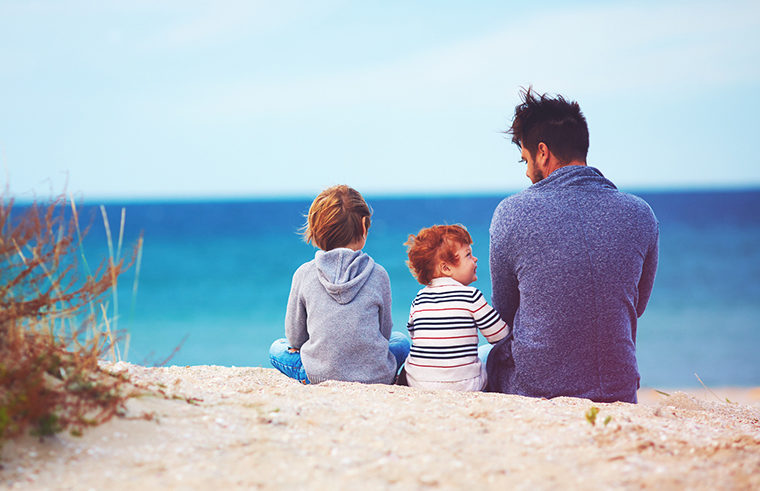 Father sitting on sand at beach with two children - feature