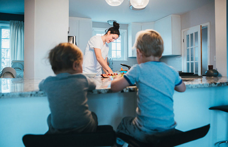 Two boys sitting on stools at kitchen bench with mother cooking - feature