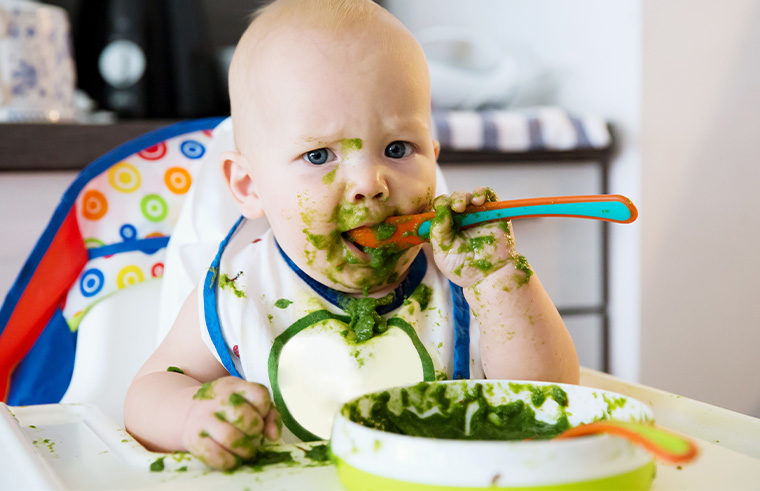 A baby eating something green and getting it all over him