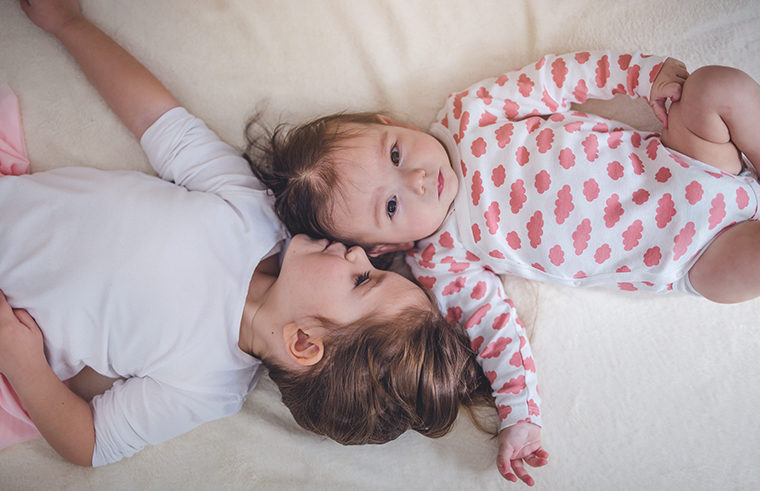 Baby and toddler sisters lying on bed overhead - feature