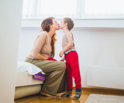 Mother sitting on bed in bedroom kissing son - feature