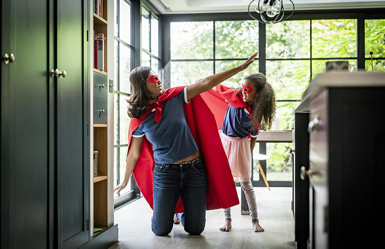 Mother playing with girl in kitchen wearing superhero capes - feature